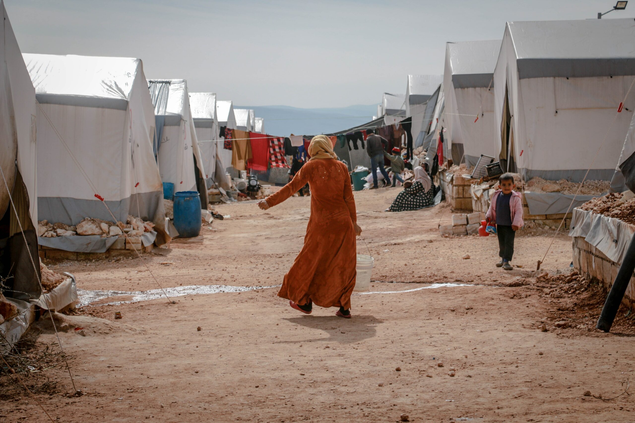 Woman carrying bucket on dirt road between tents in a refugee camp.