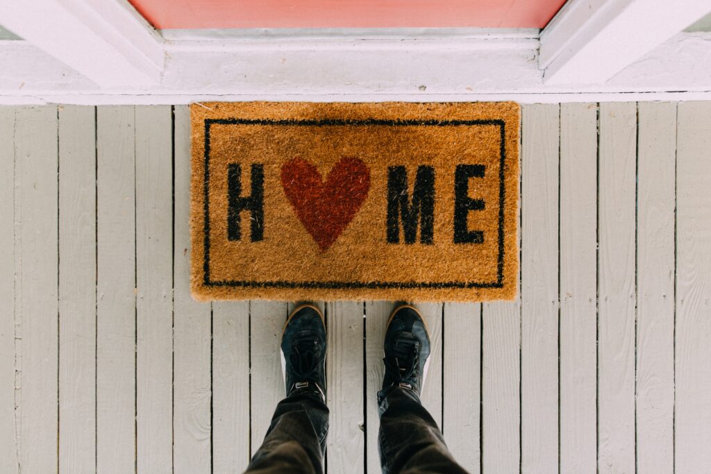 A person's feet, standing in front of a doormat with the word HOME written on it, and the 'O' replaced with a red heart.