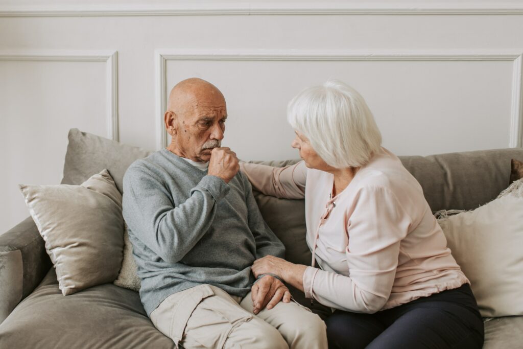 Elderly man sitting on couch holding hand to his mouth to cover a cough. A woman is sitting next to him trying to give comfort.